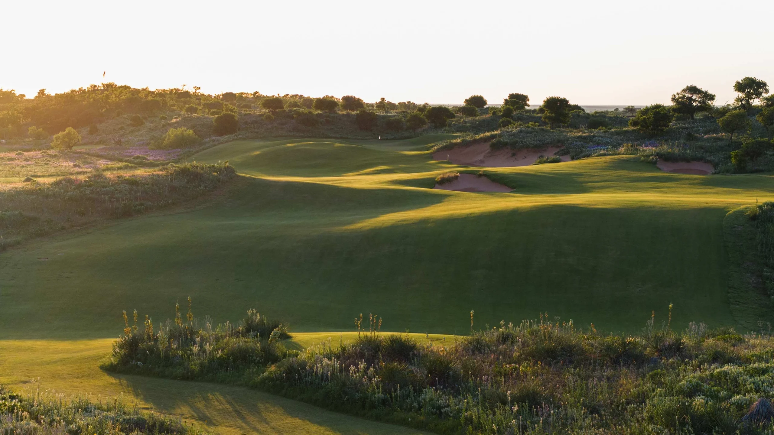 Scenic golf course at sunset with rolling green fairways, sand bunkers, and natural vegetation under warm golden light.